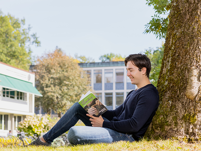 En student sitter i gresset og leser en bok utenfor Blindernveien 11, IAKH. Foto.