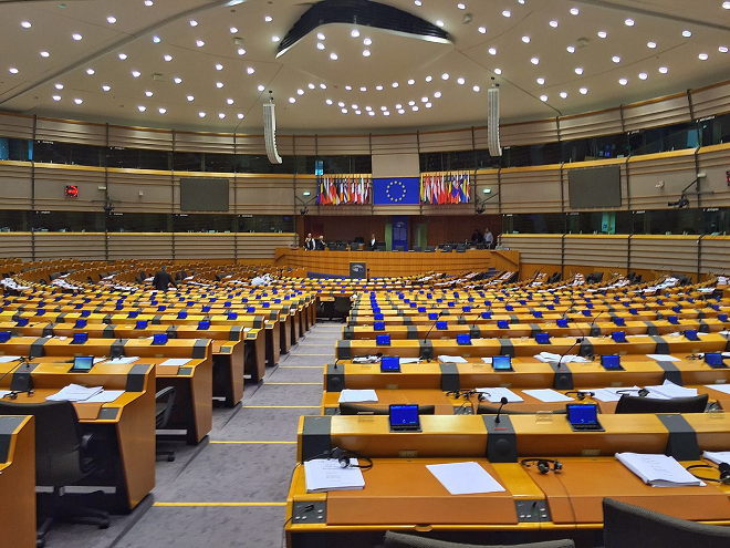 An auditorium with benches and chairs. In the background: the EU flag. Photo. 