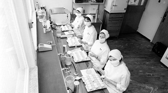 Svart hvitt bilde av fire kvinner med hvite klær og munnbind i et laboratorium. 