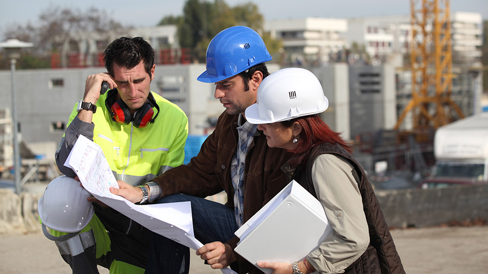 Two male construction workers and one woman wearing a helmet looks at a map or a document.