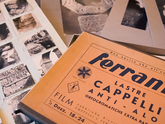 På et bord ligger en brun eldre boks for fotografier hvor det står på italiensk «Ferrania. Lastre Cappeli». Rundt ligger sort-hvitt fotografier.