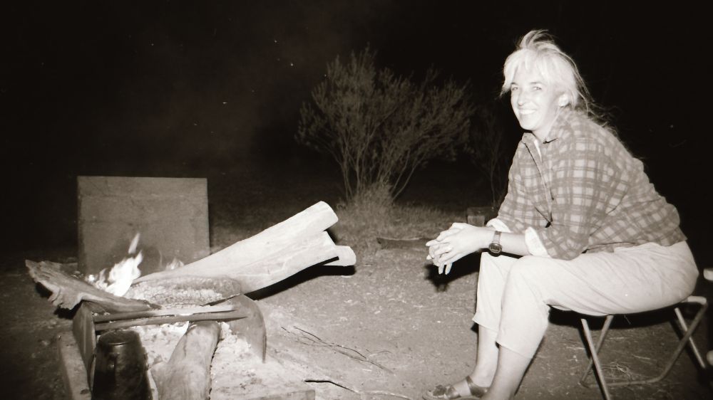 A black and white photo of the anthropologist Deborah Bird Rose sitting by a fireplace.