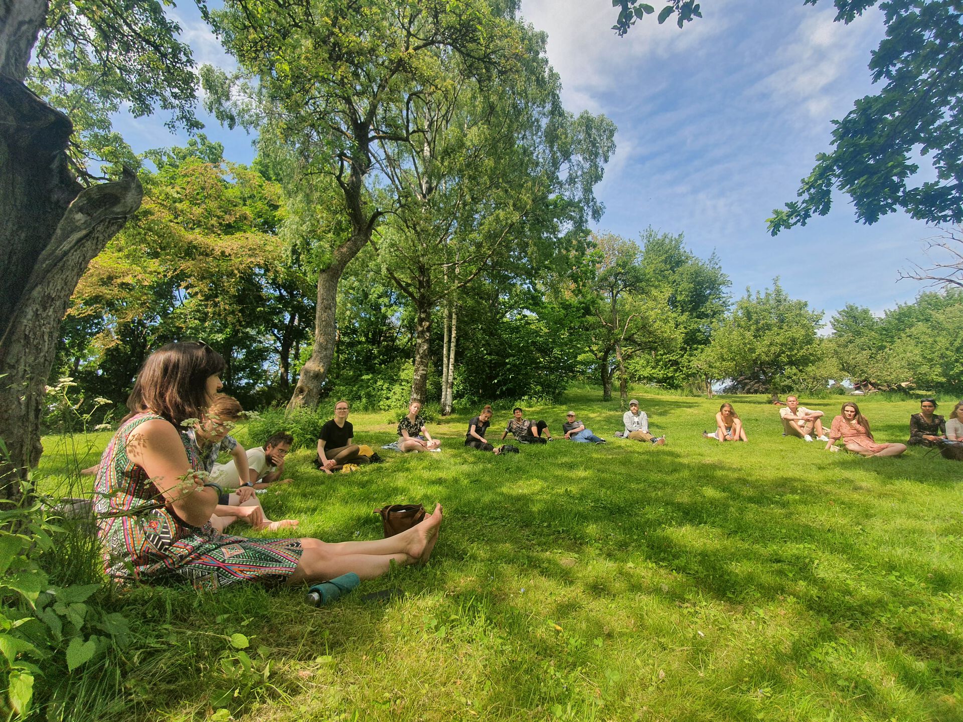 Picture of students meditating in a field of green grass. Lush, green trees in the background. 