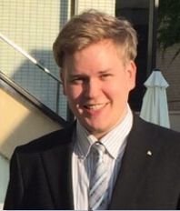 Portrait photo, young man, blonde hair, big smile, black suit, white shirt, blue tie, outside, sun, yellow wall in the background