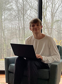 Portrait photo, young man, blonde hair, short hair, smile, white sweater, black trousers, sitting in a black comfortable chair, indoors, windows showing trees in the background