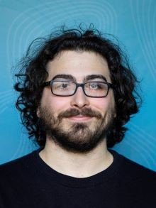 portrait photo of smiling man, blue background, black curly hair,beard and mustache, glasses with black frames, black sweather
