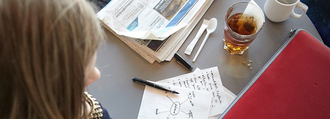 Hodet på en person bakfra, som ser ned på et papir med skriblerier, og både et glass med te og en kopp kaffe, og en avis og et dokument. Foto.