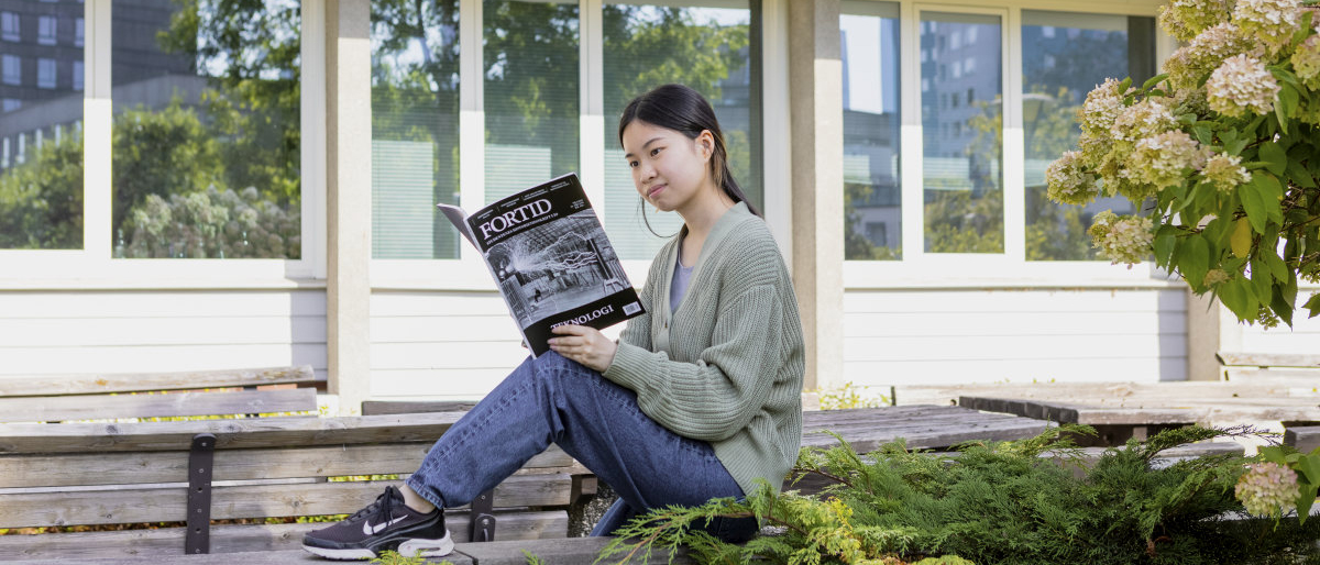 Student som sitter på en benk utenfor IAKH og leser studenttidsskriftet Fortid. Foto.