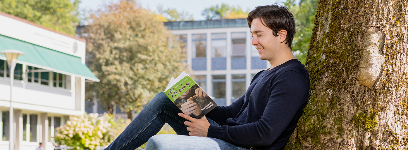 Male student sitting on the lawn reading a book, behind him you can see Blindernveien 11, IAKH. Photo.
