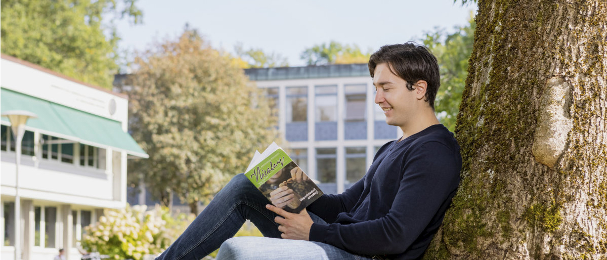 Male student sitting on the lawn reading a book, behind him you can see Blindernveien 11, IAKH. Photo.