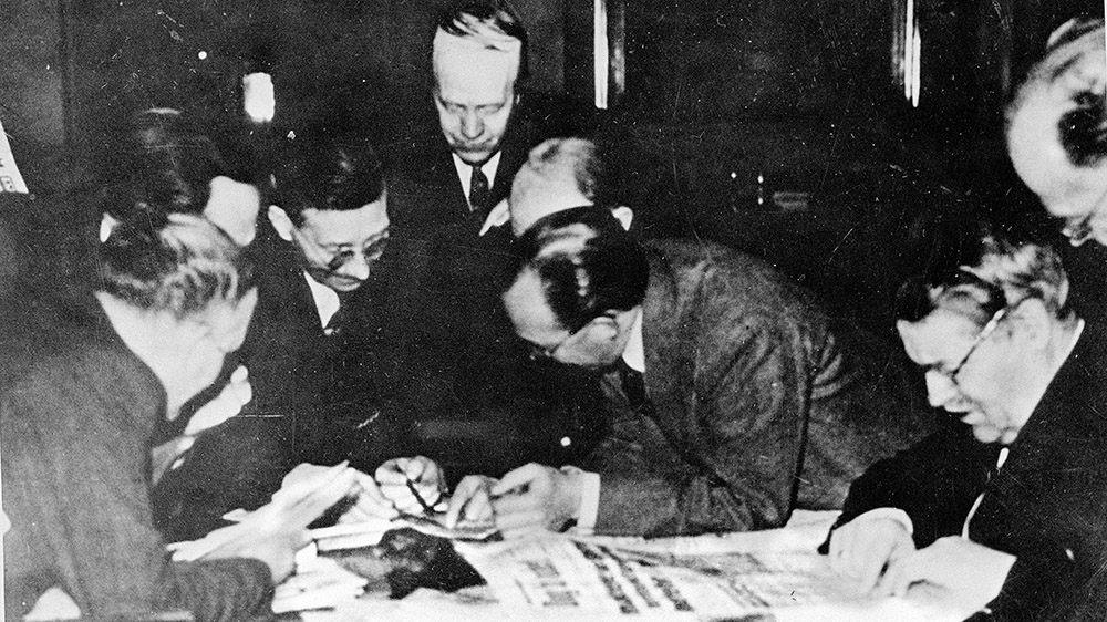A group of men in suites leaning over a table with newspapers. Black and white photo.