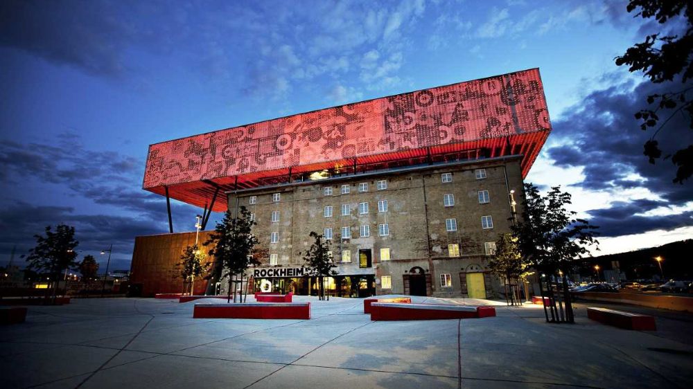 Photo of of Rockheim's facade against a moody sky. Rockheim is a brown concrete building and has a large red light installation which covers the entire roof. In front of the building there are benches and common area.