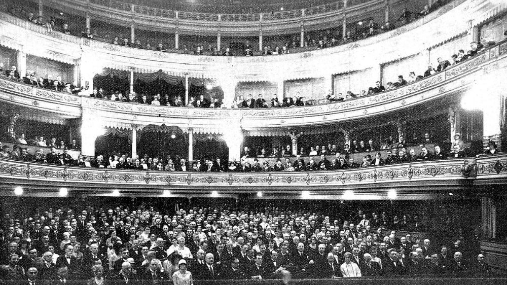 A black and white photograph of a theatre audience, taken from the vantage point of the stage