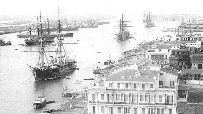 Several large sailing ships are stationary in a canal. Several large buildings and a long pier line the canal. Black and white photo.