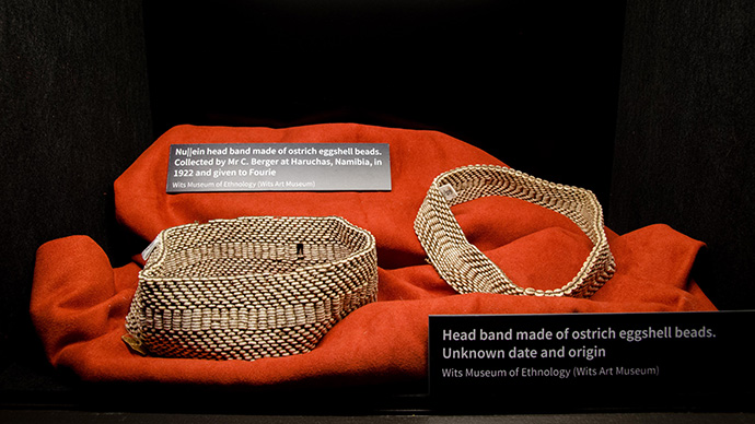 Two headbands made of beads from ostrich eggshell. They are placed on a red cushion. Photograph.