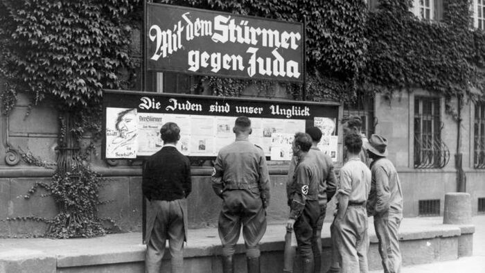 Bildet er i sort hvitt og viser en gruppe hvite menn som er samlet rundt en oppslagstavle. De har ryggen mot kamera. Tavlen viser antisemittisk oppslagstavle. 