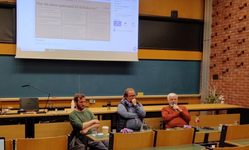Tre mennesker er avbildet sittende ved en pult i et auditorium. I bakgrunn vises en hvit skjerm med tekst.