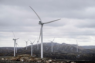 Bildet viser en vindmøllepark i åpent landskap. Det er en overskyet dag og det er fjell i bakgrunnen. Vindmøllene er hvite og himmelen er grå. 