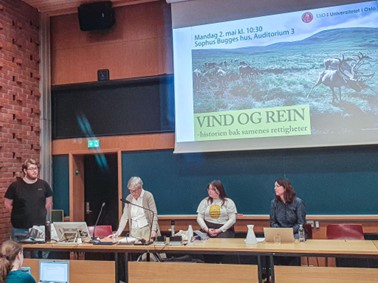 Paneldeltagerne står bak en pult i et auditorium. Bak panelistene er en tavle samt en skjerm som viser et bilde av en reinsdyrflokk over en sommervidde. Bildet inneholder informasjon om arrangementet Vind og rein.
