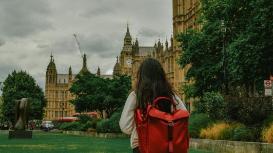 Person med mørkt langt hår og rød ryggsekk står i en park og ser mot Westminster-palasset i London. Overskyet himmel. Foto