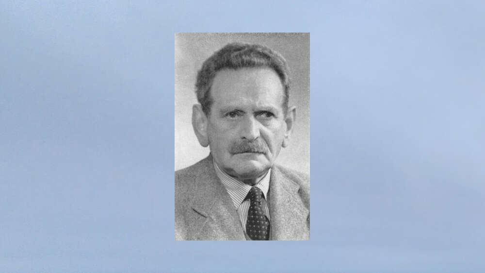 A black and white photo of a man with a shirt, tie and a jacket. The background is blue/grey.