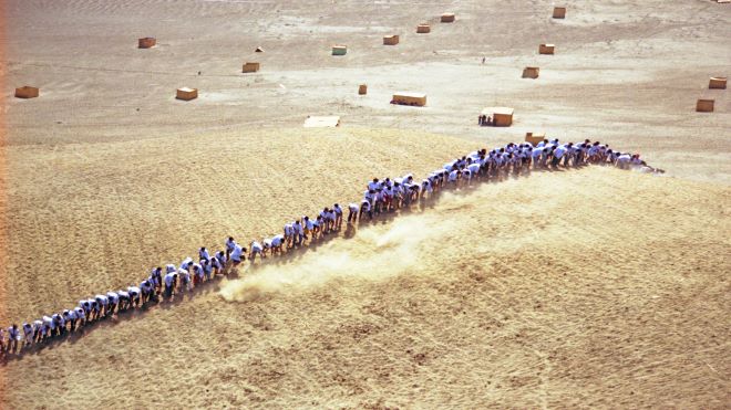People in white shirts standing in a row in a desert, shovelling sand. Photo.