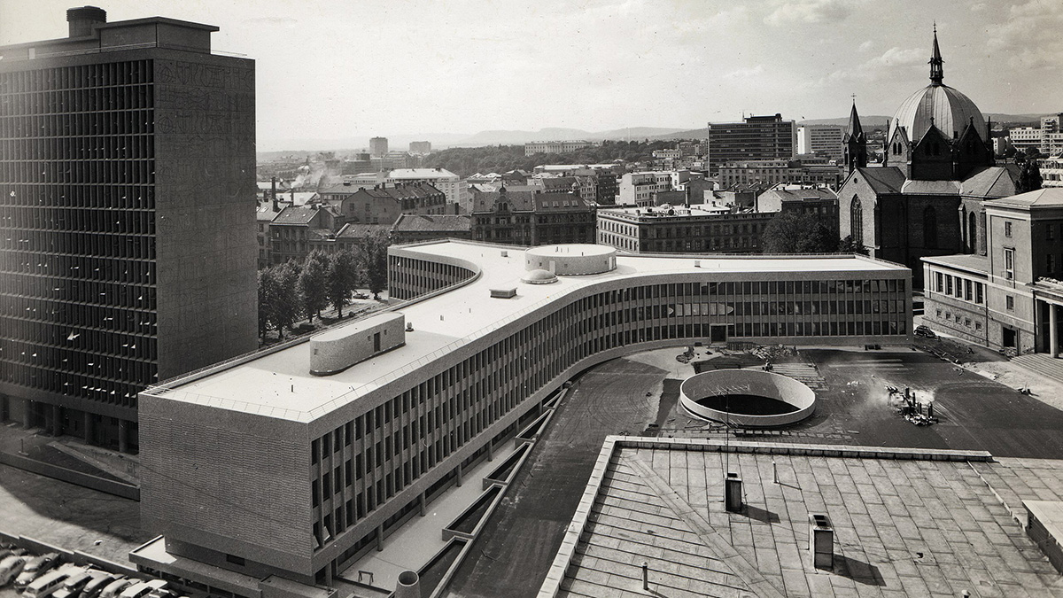 Svart-hvitt foto av regjeringskvartalet. En høy blokk i venstre kant av bilde, en y-formet lavblokk i sentrum av bildet. I bakgrunnen ser man byen.