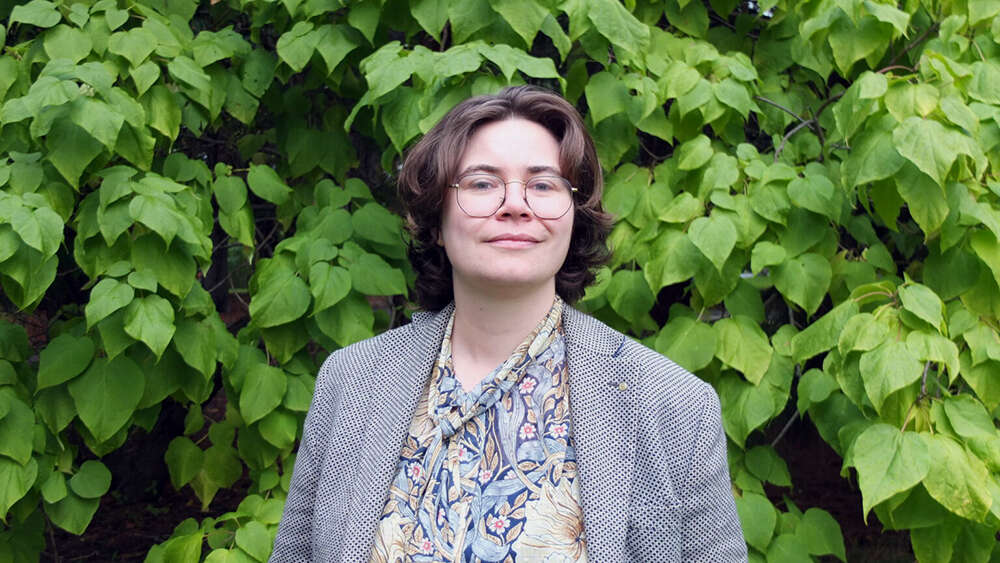 En dame rundt 40 år med briller, mørkt hår og grå dressjakke foran en grønn bakgrunn.