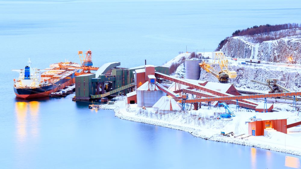Large container cargo ship docked at the cargo pier. On the pier, there are several large installations for container loading, and the ground around the pier is covered in snow. Photo.