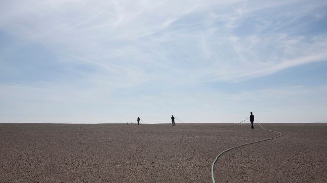 Landskap med sand og himmel, tre personer som bærer et tau. Fotografi.  