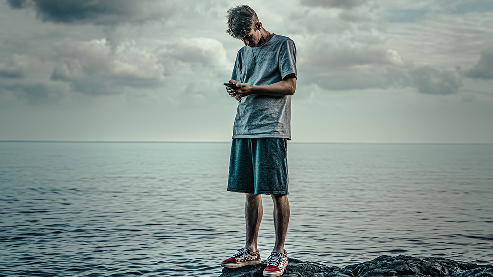 En ung mann ser på en mobiltelefon mens står på en stein ved sjøen.