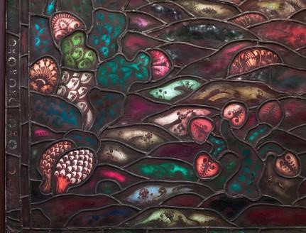 Nærbilde av et glassmaleri i ulike farger med blader og noe som ser ut som kongler. Foto.
