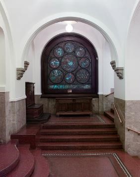 Innendørsbilde fra en murbygning med en trapp med fire trinn, hvitt tak og tykke buer. Innerst i rommet et glassmaleri med sirkler. Foto.