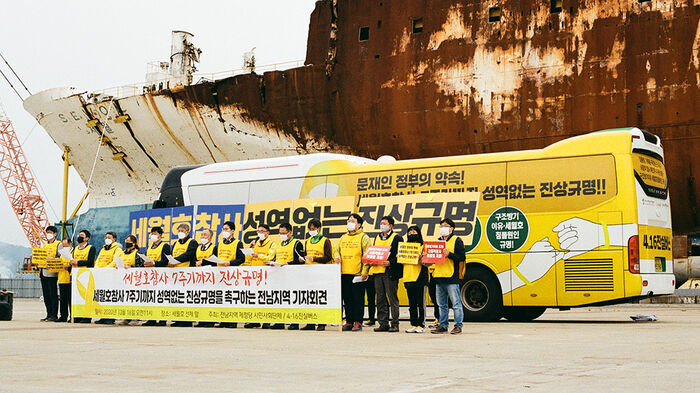 A group of people with a banner standing in front of a bus. In the background, a large ship. Photo. 