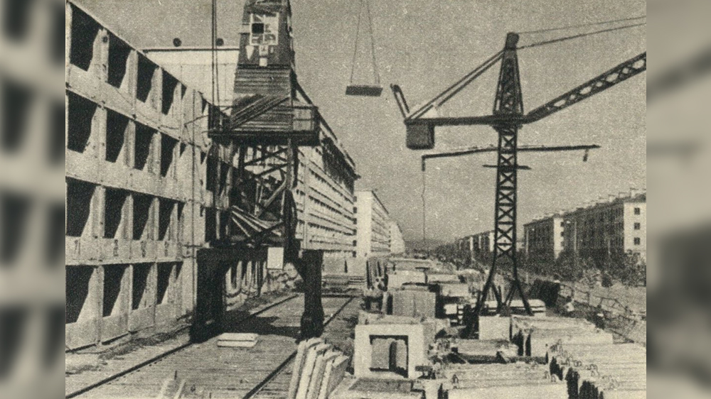A black and white photo of a building site. Buildings in the background with two hoisting cranes in the front. Photo 