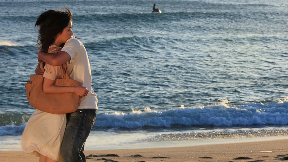 Image may contain a man and a woman hugging on the beach next to the ocean.