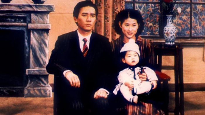 Image contains a man in a suit, a woman in a black dress and a baby in a white cap and a full-body suit.