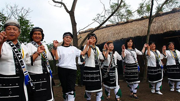 The image shows women wearing white shirts and black skirts with lines of grey, all holding hands and dancing in their village. 