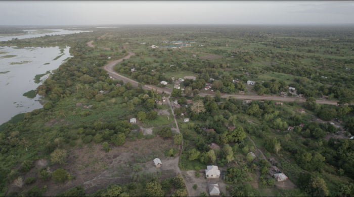 Aerial view of a rural landscape with scattered houses, winding roads, and lush greenery, bordered by a river on the left side.