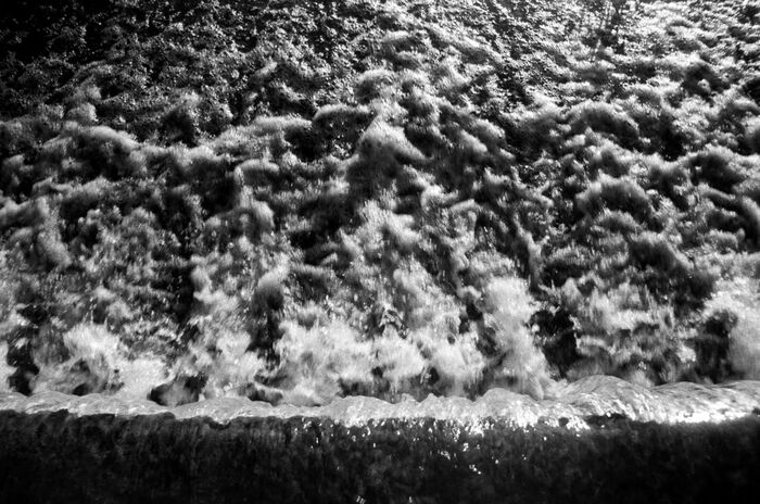 Black and white image of turbulent water cascading over a ledge, resembling a waterfall or dam overflow.