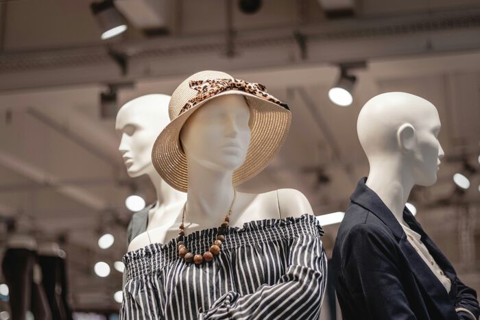 Mannequins in a clothing store display wearing fashionable outfits, including a striped off-shoulder top, necklace, and a sunhat with a bow.