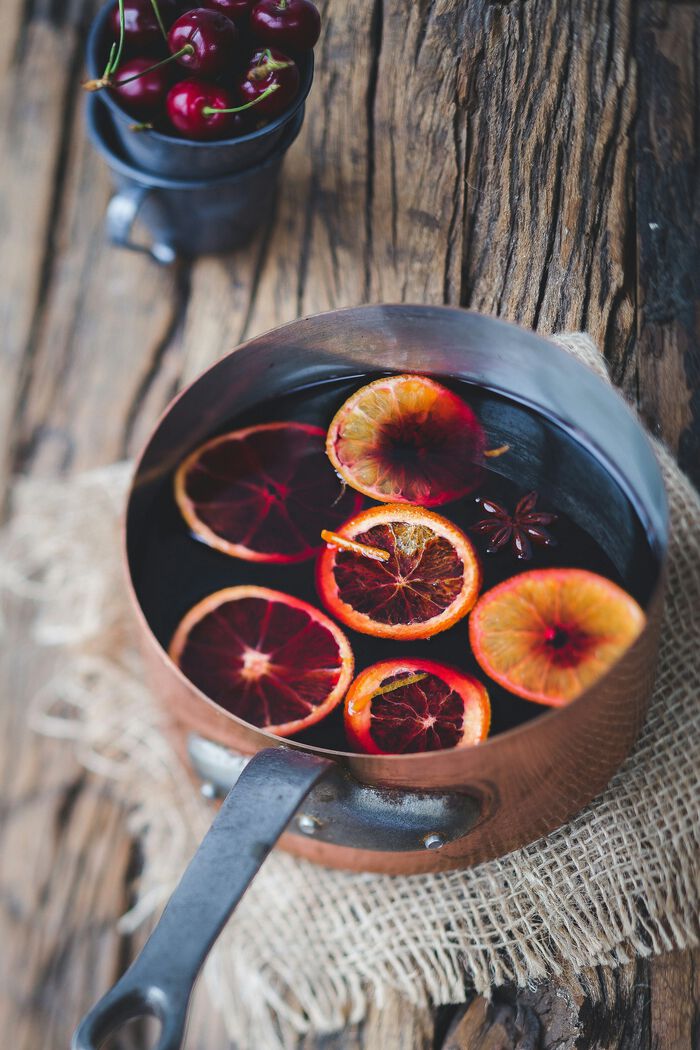 Copper saucepan with mulled wine, garnished with slices of blood orange and star anise, set on a rustic wooden surface with a cup of cherries in the background.
