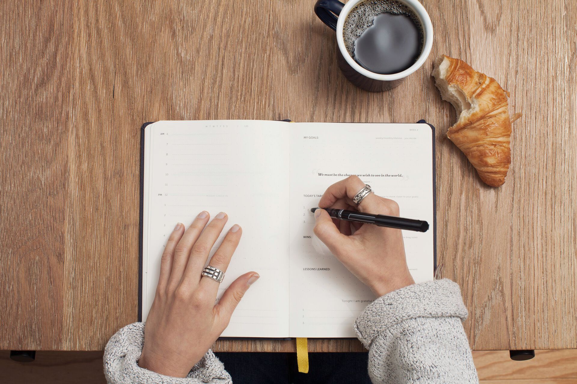 Person writing in an open notebook on a wooden table with a cup of coffee and a croissant nearby.