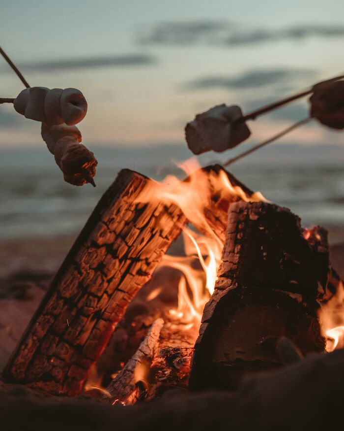 Close-up of a campfire at dusk with bread and marshmallows roasting on sticks, set against a blurred lake and cloudy sky.