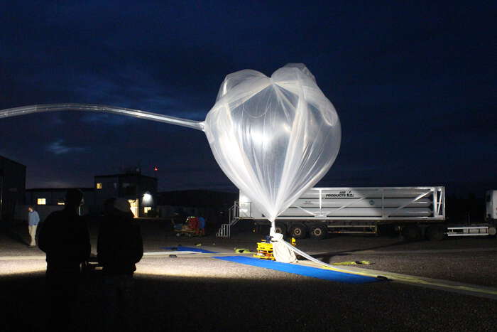 A large scientific balloon being inflated outdoors at night, illuminated by bright lights, with silhouetted people and equipment nearby.