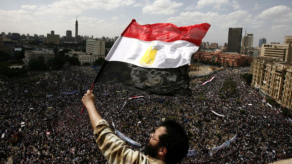Mann vifter med egyptisk flagg med demonstrasjoner ved Tahrir-plassen i bakgrunnen. 