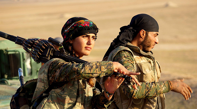 Kurdish YPG-soldiers in Rojava. A woman with military gear and weapon, a man in the background.