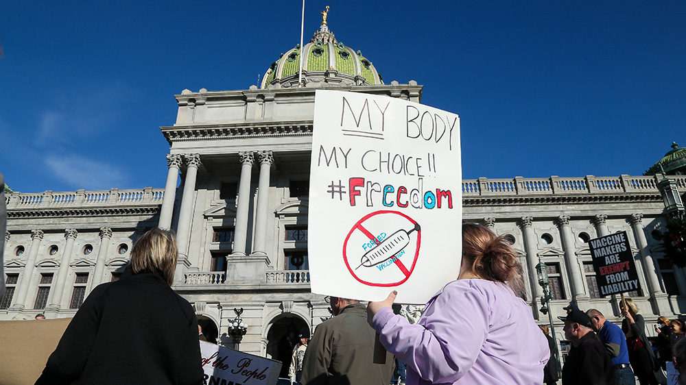 A group of people protesting in front of big building. A woman wearing a purple sweater hold a poster saying "My body. My choice. #freedom." and a cross over a drawing of a syringe and the words "fored vaccination".