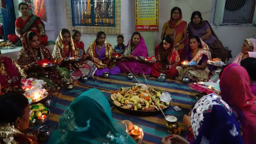About 20 colourfully dressed women are seated in a circle in a temple room. Photo.