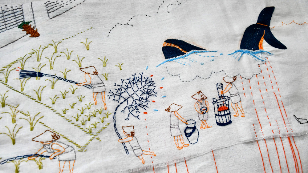 Embroidery on white textile. One sees a whale, humans and grass embroidered in different colours.
